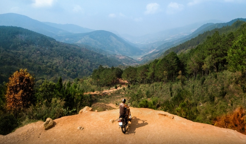 Adventurer enjoy touring motorbike stopped by a cliff viewpoint on mountain peak with pine forest in Vietnam