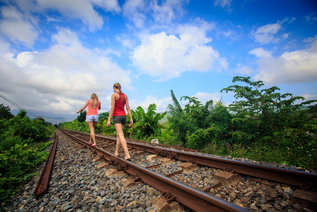 two young girls in shorts walk along railway past plants 